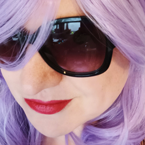 A close up of the face of a white person in a purple wig and sunglasses, wearing red lipstick and smirking.
